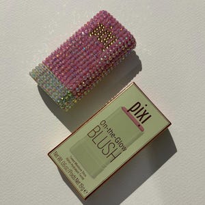 May include: A pink blush stick and its box. The blush stick is covered in pink rhinestones and gold lettering. The box is light green with the words "pixi On-the-Glow BLUSH" and the product's weight.