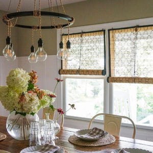 May include: A circular black metal chandelier with hanging Edison bulbs suspended by tan rope cords. The fixture features a round frame wrapped in rope, illuminating a dining table set with clear glass tumblers, plates, and a large glass vase.