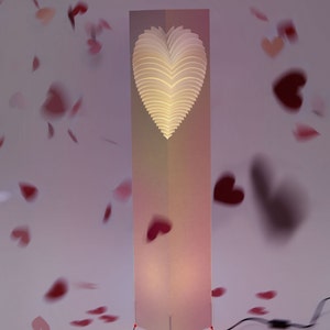 May include: A white heart-shaped lamp with a layered design, standing on a pink and white base with a cord and plug. The lamp is surrounded by pink hearts.