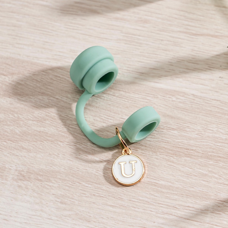 May include: A light green silicone bottle cap with a gold-colored charm. The charm is a white circle with the letter "U" in the center. The cap is on a light wood surface.