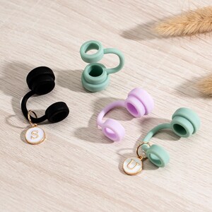 May include: An assortment of colorful silicone bottle stoppers. The stoppers are in black, light green, and lavender. Each stopper has a loop and a small gold charm with a letter. The letters are "S" and "U".