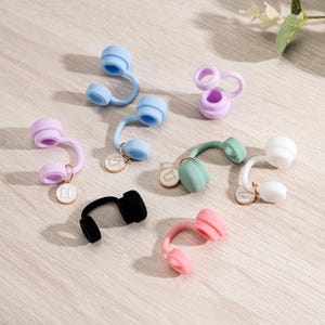 May include: A collection of colorful, small, headphone-shaped accessories in various colors, including blue, pink, green, white, and black. Each accessory has a curved stem and a circular charm with a letter. The items are arranged on a light-colored surface.