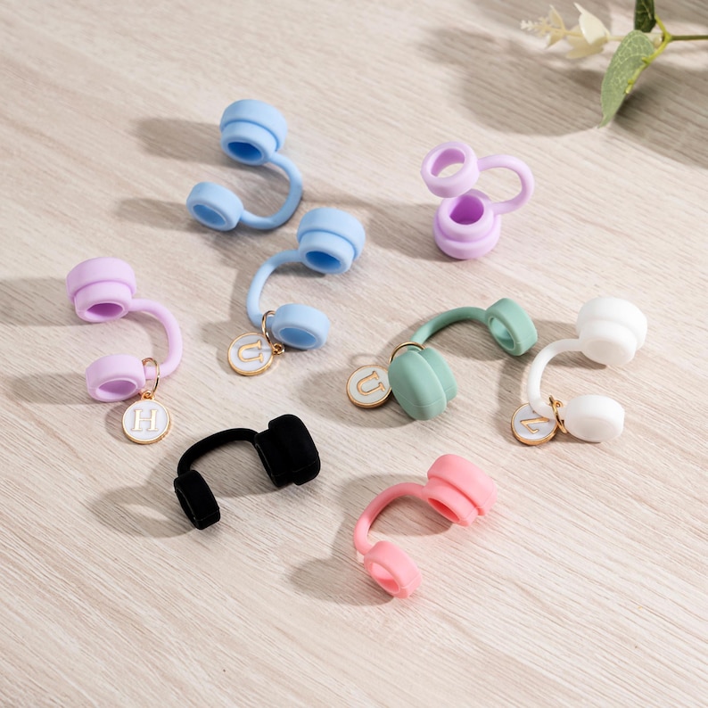 May include: A collection of colorful, small, headphone-shaped accessories in various colors, including blue, pink, green, white, and black. Each accessory has a curved stem and a circular charm with a letter. The items are arranged on a light-colored surface.