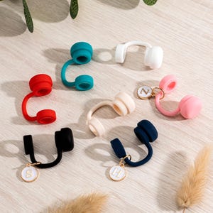 May include: A collection of colorful, silicone wine bottle stoppers in various hues, including red, teal, white, pink, beige, black, and navy. Some stoppers feature a small, gold-colored charm with a letter. The stoppers are arranged on a light-colored wooden surface.