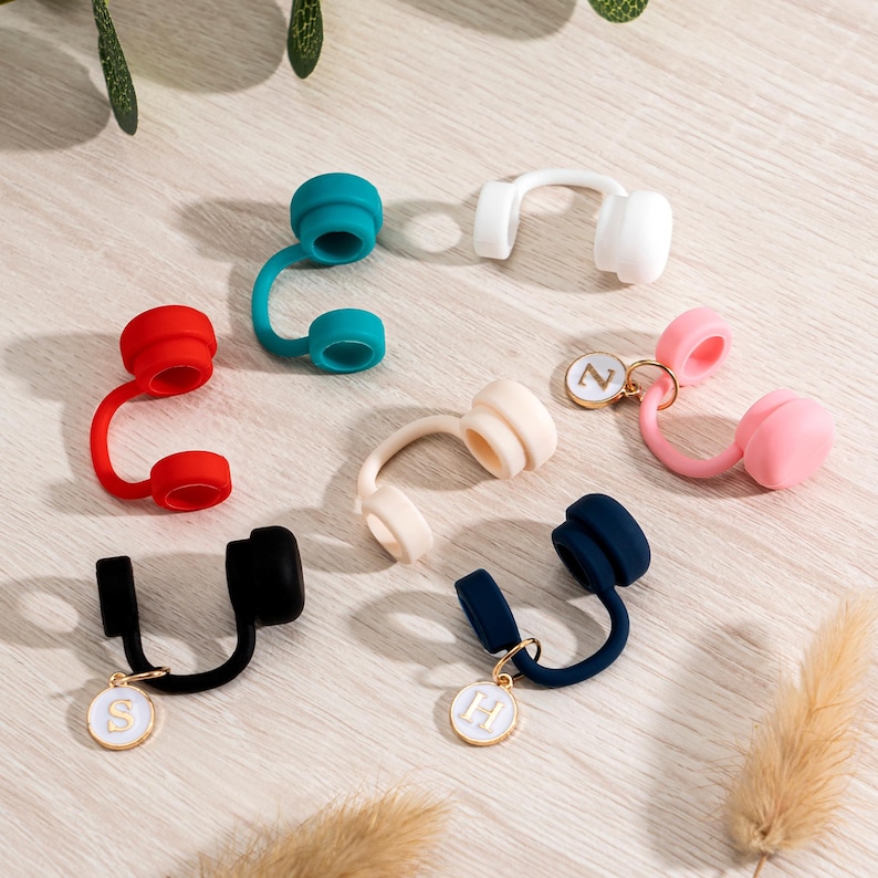 May include: A collection of colorful, silicone wine bottle stoppers in various hues, including red, teal, white, pink, beige, black, and navy. Some stoppers feature a small, gold-colored charm with a letter. The stoppers are arranged on a light-colored wooden surface.