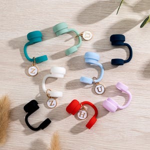 May include: A collection of colorful silicone straw toppers in various shades, including teal, mint green, white, light blue, navy, black, red, and lavender. Each topper has a gold-colored charm with a letter. The toppers are arranged on a light wood-grain surface.