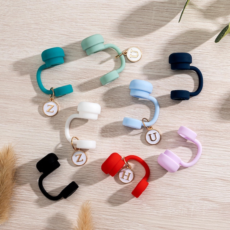 May include: A collection of colorful silicone straw toppers in various shades, including teal, mint green, white, light blue, navy, black, red, and lavender. Each topper has a gold-colored charm with a letter. The toppers are arranged on a light wood-grain surface.
