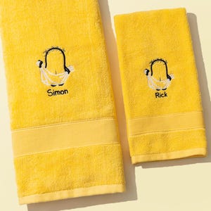 Könnte beinhalten: Zwei gelbe Badetücher mit gestickten Cartoonfiguren. Auf einem Handtuch steht der Name "Simon" unter der Figur, auf dem anderen "Rick". Die Handtücher haben ein dekoratives Band am unteren Rand.