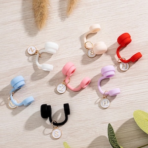 May include: A collection of colorful, silicone earbud holders in various colors, including white, pink, blue, red, black, and lavender. Each holder features a gold-colored charm with a letter. The holders are designed to wrap around the earbud cord.