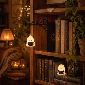 May include: A white ghost-shaped light fixture hanging from a black metal scrollwork wall bracket, and a matching white ghost-shaped light sitting on a wooden bookshelf. Both ghost figures feature black eyes and hold a small black open book.