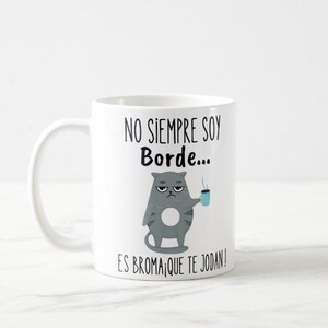 Puede incluir: Taza de cerámica blanca con asa negra, con una ilustración de un gato gris gruñón sosteniendo una taza azul. La taza tiene el texto negro "NO SIEMPRE SOY Borde... ES BROMA! QUE TE JODAN!"