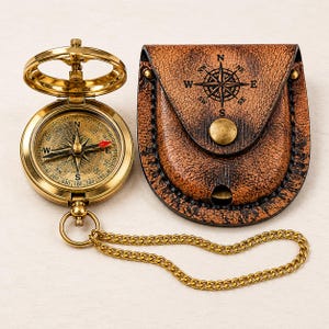 May include: A polished brass pocket compass with a hinged lid and a matching metal chain, positioned next to a brown textured leather pouch featuring a stamped black compass rose design on the flap and a central brass snap button closure.