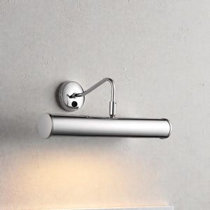 Op de afbeelding: Een gepolijste chromen schilderijlamp. De lamp heeft een ronde wandbevestiging, een gebogen arm en een cilindrische lichtkop. De armatuur is ontworpen om kunstwerken te verlichten en is gemonteerd op een getextureerde, lichtgekleurde muur.