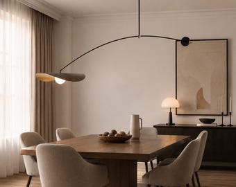 May include: A black metal pendant light with a long, curved arm and a single glowing bulb housed under a wide, flat, dark-toned shade. The fixture is suspended from the ceiling by a thin rod, featuring a minimalist design with brass accents.