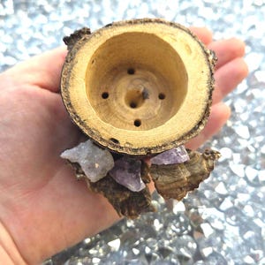 May include: A small, circular wooden planter or holder crafted from a natural log slice with visible bark on the exterior. The interior is hollowed out with four small drainage holes at the base. It is decorated with raw amethyst crystals.