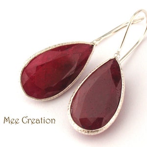 May include: Teardrop earrings featuring a deep red gemstone set in silver-coloured metal. The earrings have a long, curved hook design. The brand name "Mee Creation" is printed in brown script.