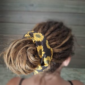 May include: A black scrunchie with a yellow sunflower pattern wrapped around a bun of light brown hair. The scrunchie is made of a silky material and adds a pop of color to the hairstyle. The background is a blurred wooden wall.