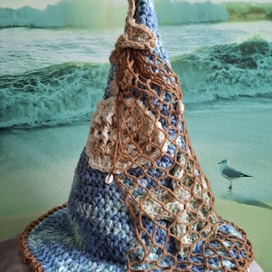 May include: A conical crochet hat featuring a variegated blue and white yarn pattern. The hat is adorned with a brown crocheted fishing net overlay, several small beige seashells, and a textured, light-colored crocheted starfish accent.
