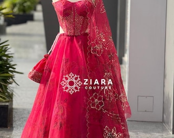 Puede incluir: Un vibrante vestido de noche rojo con un chal a juego y un pequeño bolso, exhibido en un maniquí. El corpiño está adornado con intrincados abalorios, y la falda presenta un diseño fluido. El chal está adornado con motivos florales dorados. El texto "ZIARA COUTURE" es visible.