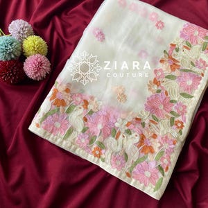 Może przedstawiać: Biały materiał tekstylny z haftem kwiatowym w odcieniach różu, pomarańczy i zieleni. Tekstylia opatrzone marką "ZIARA COUTURE". Prezentowane na głębokim czerwonym tle.