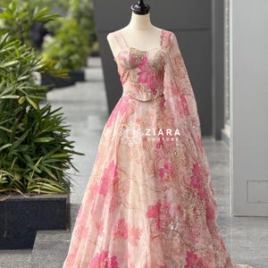 Peut inclure: Une robe de soirée sur un mannequin, ornée d'un motif floral rose sur fond clair. La robe comprend un corsage ajusté, une jupe fluide et un châle assorti. Le texte "ZIARA COUTURE" est visible sur la robe.