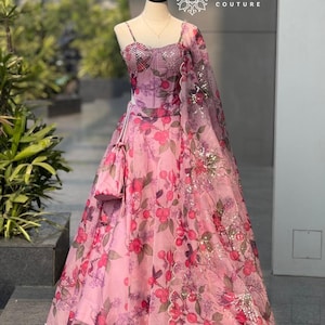 Könnte beinhalten: Ein rosa Kleid mit floralem Muster, passender Handtasche und Schal. Das Kleid hat ein eng anliegendes Oberteil mit funkelnden Verzierungen und Spaghettiträgern. Das Blumenmuster umfasst rote und violette Blumen und grüne Blätter. Der Schal ist über der Schulter drapiert.