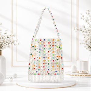 May include: A white handbag featuring vertical rows of colorful beads and clear crystals, accented with white floral appliqués. The bag includes a single shoulder strap adorned with matching beads and a bottom edge finished with long white fringe.