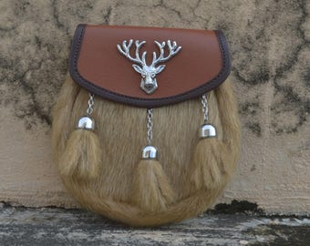 May include: A tan fur sporran with a brown leather top and a silver stag emblem. The sporran features three decorative tassels with silver caps and chain attachments. The background is a textured, neutral-toned surface.