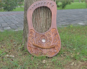 May include: A wooden lyre with a carved floral design. The instrument has a rounded body with a central sound hole and multiple strings. It is leaning against a tree trunk, with grass in the foreground.