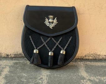 Puede incluir: Sporran de cuero negro con un emblema de cardo plateado en la solapa. El sporran tiene un cuerpo de piel y tres borlas negras colgando de cadenas plateadas. Un accesorio escocés tradicional.