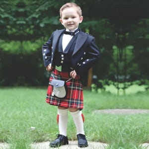 May include: A young boy in a traditional Scottish outfit. He wears a black jacket, vest, and a red and green plaid kilt. The outfit includes a black bow tie, white knee socks, and black shoes. A small grey sporran hangs from his kilt.