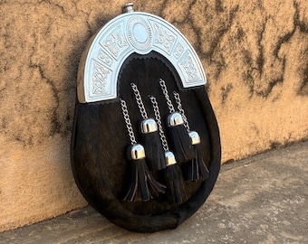 May include: A black leather sporran with a silver cantle featuring Celtic knot designs. The sporran has a fur body, silver chain tassels, and a decorative silver top. The tassels are black with silver caps.