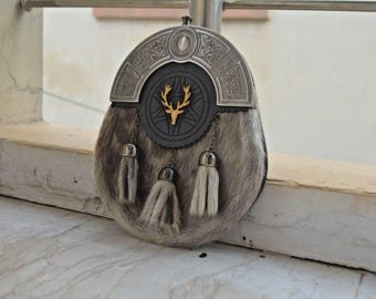 May include: A Scottish sporran with a silver cantle and a fur body. The cantle has Celtic knotwork and a central emblem with a gold stag. Three tassels hang from the bottom, attached by chains.