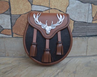 May include: A brown leather sporran with a silver stag head emblem. The sporran features a black front panel and three brown tassels. The top flap is brown with a decorative silver stag head.