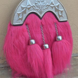 Puede incluir: Un sporran rosa intenso con un cantle plateado. El sporran tiene una textura esponjosa y tres borlas con tapa plateada colgando de cadenas. El cantle est&aacute; decorado con dise&ntilde;os en relieve.