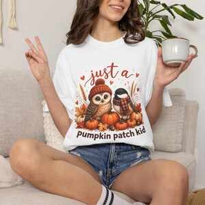May include: A white t-shirt featuring a graphic of an owl wearing a knit hat and a bird wearing a scarf, surrounded by pumpkins, autumn leaves, and wheat stalks. The text "just a pumpkin patch kid" is printed in brown lettering above and below.