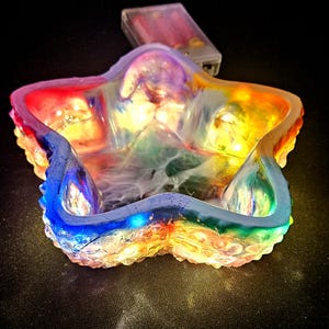 May include: A star-shaped dish made of resin, featuring a vibrant array of colours and embedded LED lights. The colour scheme includes red, orange, yellow, green, and blue. A small battery pack is visible, providing power to the lights.