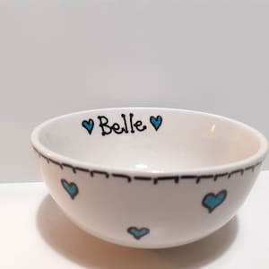 May include: White ceramic bowl with a hand-drawn design. The bowl features the name "Belle" in black script with a teal heart above it. Teal hearts are also scattered around the bowl. A black dashed line borders the rim.