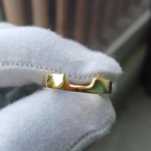 Op de afbeelding: Een gepolijste gouden ring met een gladde, doorlopende band met een opvallende U-vormige uitsparing of inkeping langs de bovenrand. Het metalen oppervlak is sterk reflecterend, met heldere accenten en een warme, metallic gele kleur.