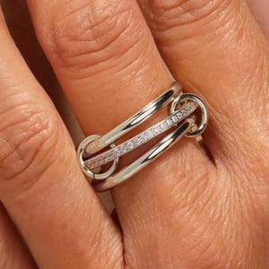 May include: A silver ring with a unique design. The ring features three interconnected bands, with one band adorned with small, sparkling stones. The ring is shown on a person's finger, highlighting its elegant and modern style.