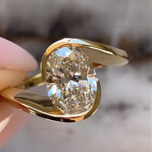 Op de afbeelding: Een gouden ring met een grote, ovaal geslepen diamant. De diamant is gezet in een bezel-zetting met een gebogen, modern design. De ring is een sieraad.