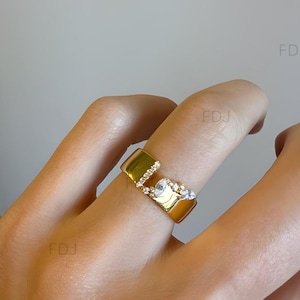 Könnte beinhalten: Ein goldfarbener, offener Ring mit einer breiten, polierten Ringschiene und einer Lücke. Eine Seite der Öffnung ist mit einer Reihe kleiner, klarer, runder Steine besetzt, während die andere Seite eine Ansammlung klarer, birnenförmiger Steine zeigt.