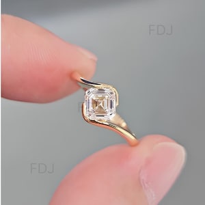Puede incluir: Un anillo de oro con una piedra preciosa transparente de corte cuadrado montada en una banda de estilo bypass. La banda se curva alrededor de la piedra central, que tiene un patrón de facetas escalonadas. Las letras FDJ son visibles al fondo.