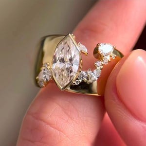 Puede incluir: Un anillo de oro con un gran diamante talla marquesa como pieza central. El anillo está adornado con diamantes más pequeños, redondos y talla marquesa, creando un diseño único y elegante. El anillo es sostenido por una persona.