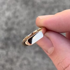 May include: A gold-colored ring with a rectangular, white-colored face. The ring is held between fingers, showcasing its polished surface and design. The ring is a piece of jewelry.