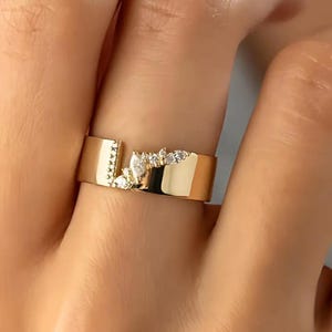 Puede incluir: Un anillo dorado con un diseño único. El anillo presenta una banda ancha con una abertura y está adornado con pequeñas piedras preciosas transparentes. Las piedras preciosas están agrupadas en un patrón decorativo, agregando brillo a la joya.