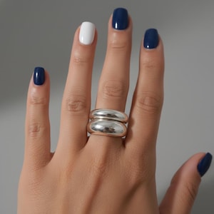Può includere: Un anello d'argento con un design a doppia fascia è indossato su una mano con smalto blu navy e bianco. L'anello ha una forma liscia e arrotondata e riflette la luce, creando un aspetto lucido. Lo sfondo è grigio neutro.