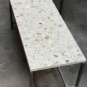 Könnte beinhalten: Ein rechteckiger Couchtisch mit einer Terrazzoplatte und einem Metallrahmen. Die Tischplatte weist ein Mosaik aus weißen, grauen und beigen Steinfragmenten auf. Der Metallrahmen hat eine dunkelgraue Oberfläche. Der Tisch ist ca. 122 cm lang.