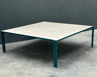 Op de afbeelding: Een vierkante salontafel met een lichtgrijs blad en teal-kleurige poten. De tafel heeft een modern design en is geschikt voor binnen of buiten. De afmetingen van de tafel zijn ongeveer 100 cm bij 100 cm.