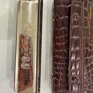 Pode incluir: Uma harmónica M. Hohner de cor prateada com texto gravado e emblemas circulares, acompanhada por um estojo de transporte castanho escuro com textura de crocodilo e o texto HOHNER-Chromonica 280 - C impresso na aba frontal.